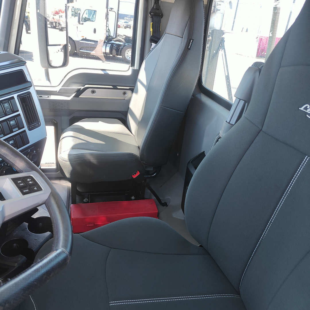 Mack Anthem truck cockpit interior showing driver seat and passenger seat with gray upholstery; red toolbox under the dash; steering wheel and dashboard visible.