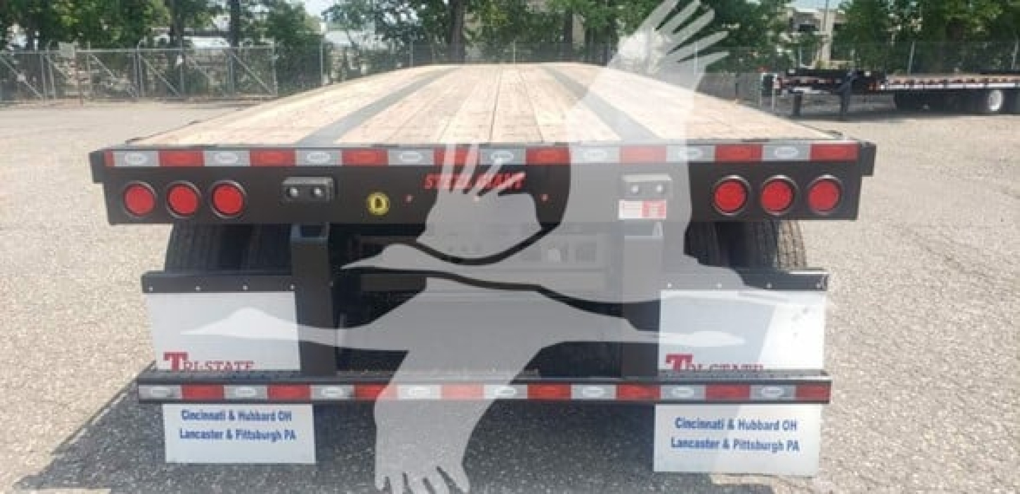 Flatbed trailer rear view with tandem axles, red rear lights, and protective mudflaps; DOT reflective tape along edges; blank wooden deck; stock and dealer labels visible.