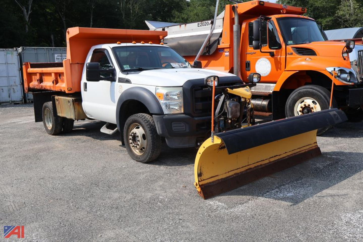 Ford F-550 dump truck with plow showing white crew cab and orange dump body in a vehicle lot with a yellow snow plow attachment in the foreground.