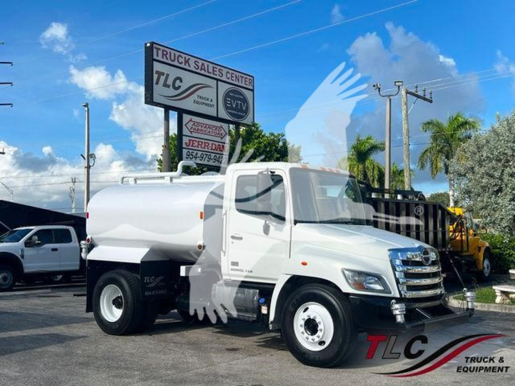 Hino 338 water truck with tank body in a dealership lot, white cab and tank, front grille, sign boards in background.