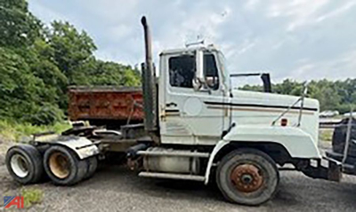 Semi tractor truck with plow in a rural lot; white cab, tall exhaust stack, flatbed trailer attachment, large front wheel and dual rear wheels; Freightliner-like design.