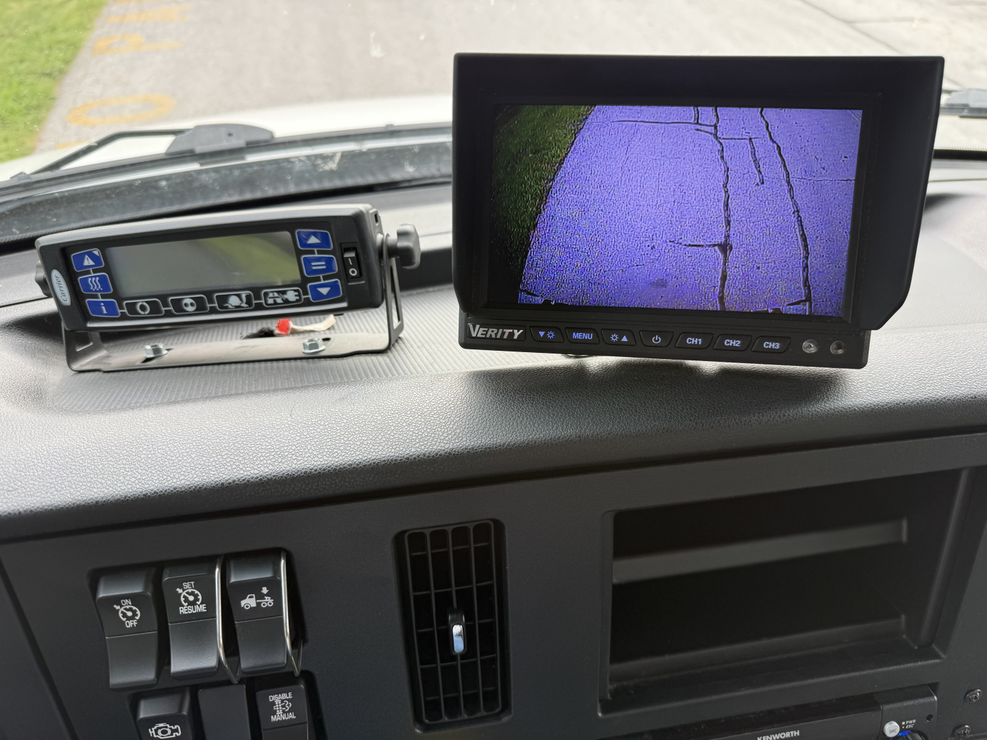 Dashboard view of a 2023 Kenworth T380 cab interior showing a digital driver monitor and a Verity screen with rearview camera display, plus control switches and empty storage bay.