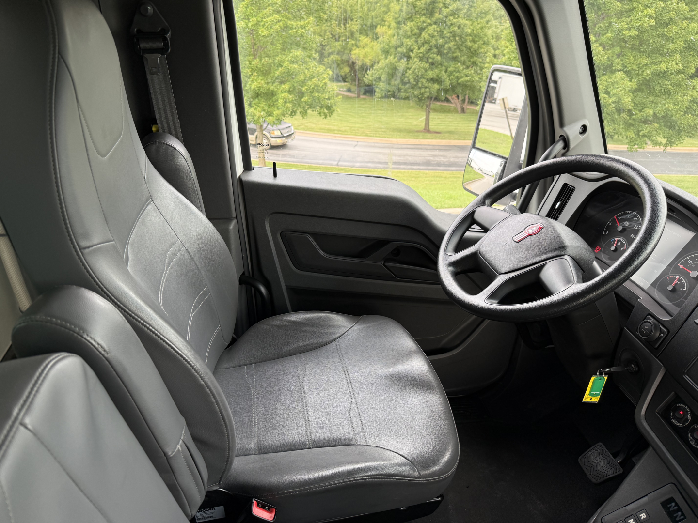 Truck cab interior of a 2023 Kenworth T380 showing driver seat and dashboard, steering wheel with Kenworth logo, gray vinyl seating, and side door panel.