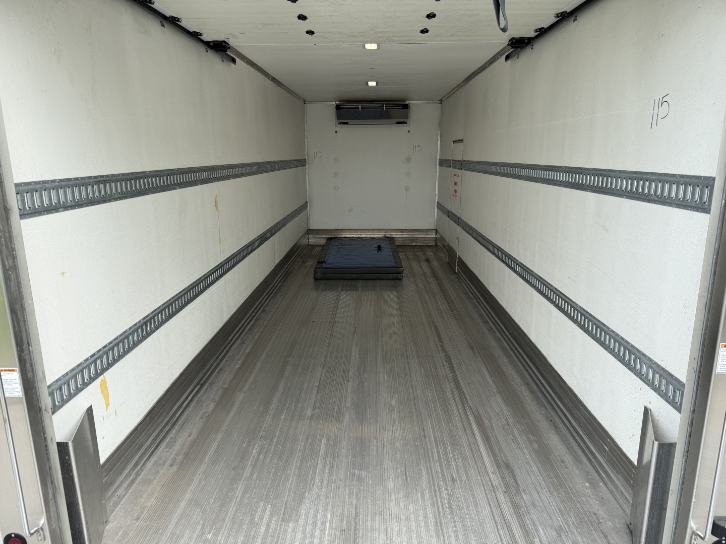 Interior of a refrigerated Kenworth truck cargo area showing empty box with aluminum E-track rails along walls, roof-mounted insulation panel, and a small pallet on the floor.