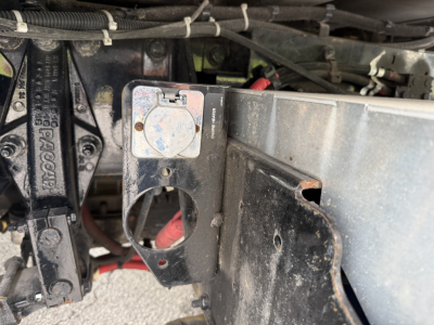 Kenworth T380 engine bay area showing a circular cap with blue center, metal mounting bracket, and multiple hoses and wiring harnesses.
