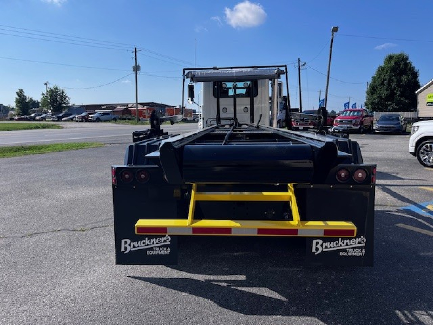 Rear view of a Mack roll-off truck with a black rear frame and yellow bumper; Buckners Equipment logos on mud flaps; red taillights; clear sky.