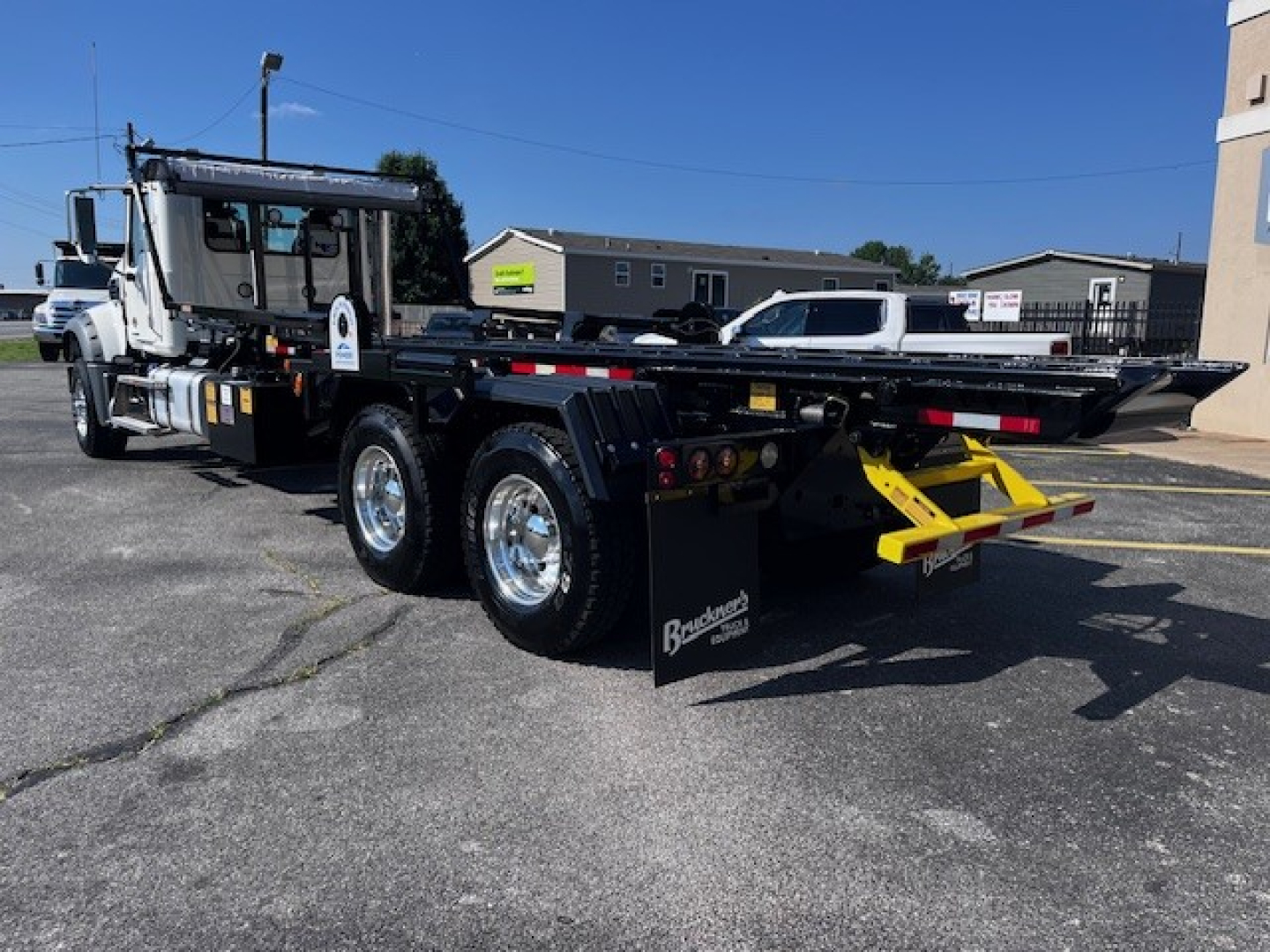 Truck rear view showing a Mack GR64F roll-off body with dual rear axles, flatbed with side rails, and bright yellow recovery arm. VIN 1M2GR3GC0TM050590 on stock number 69215.