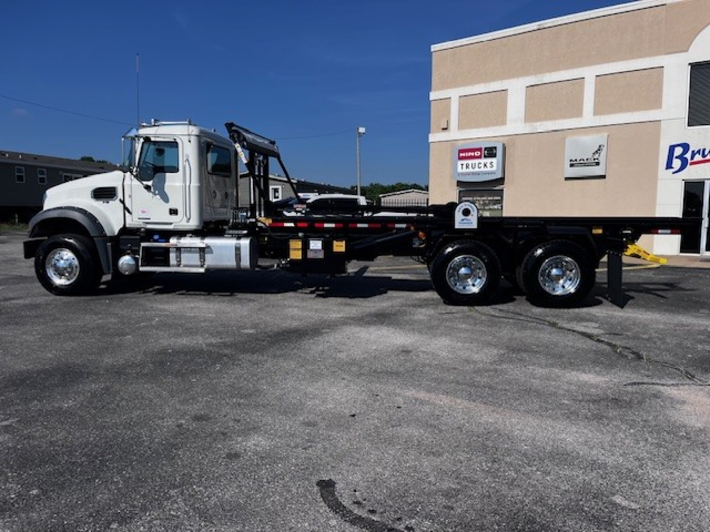 Mack GR64F road tractor with roll-off body parked in front of a building, side view showing cab, chassis, and dual rear axles.
