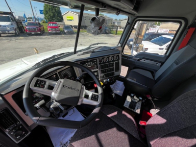 Interior view of a new 2026 Mack GR64F truck cab with steering wheel, dashboard controls, and driver seat; visible windshield and side window; some interior papers on the floor.