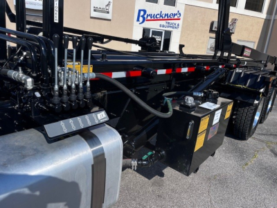 Mack GR64F truck chassis showing hydraulic lines, control levers, fuel tank, and battery/auxiliary box with labels visible.