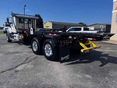 Truck rear view showing a Mack GR64F roll-off body with dual rear axles, flatbed with side rails, and bright yellow recovery arm. VIN 1M2GR3GC0TM050590 on stock number 69215.