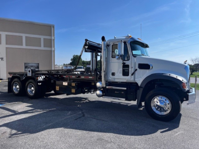 2026 Mack GR64F roll-off truck side view showing cab, chassis, and hydraulic boom with flatbed; visibility of front wheel, mirrors, and exhaust stack.