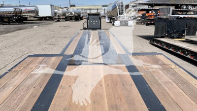 Trailer bed of a Fontaine Renegade N12 lowboy showing wooden deck planks and steel center rails with reflection of a shadowed hand,”