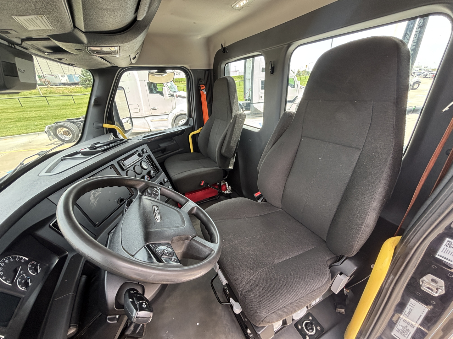 Cab interior of a Freightliner daycab showing steering wheel, driver's seat, dashboard, and passenger seat with modern control panel and gear selector visible.