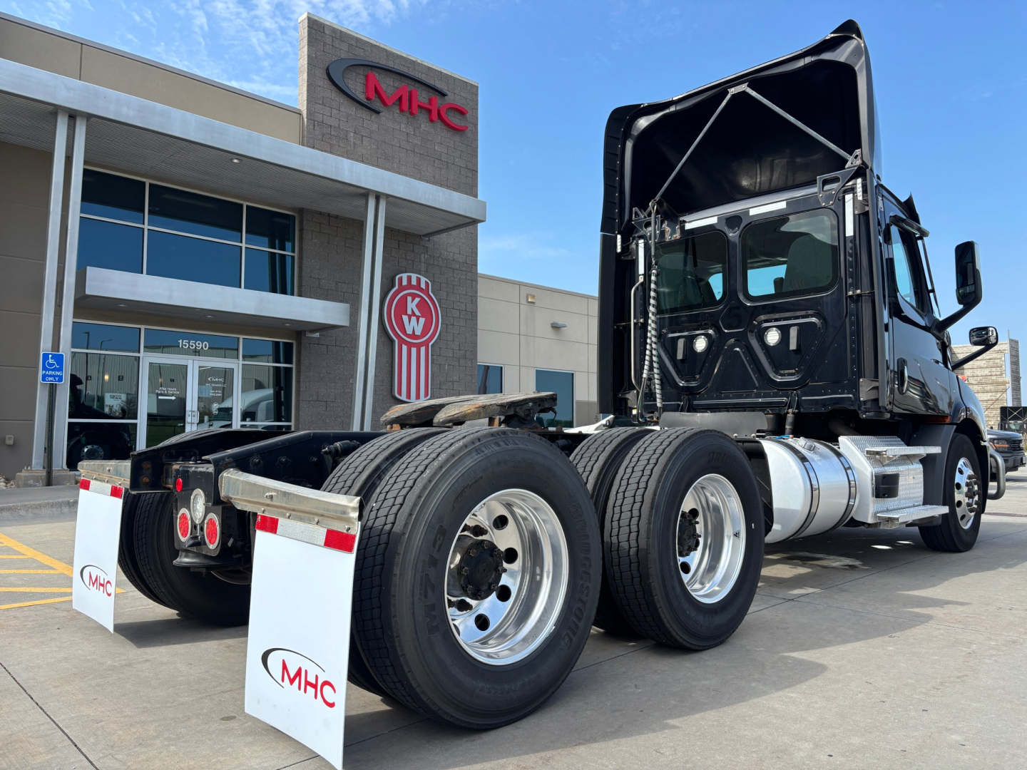 Truck chassis and cab of a Freightliner semi-truck seen from rear-left, with fifth-wheel and dual rear axles; MHC dealership building in background.