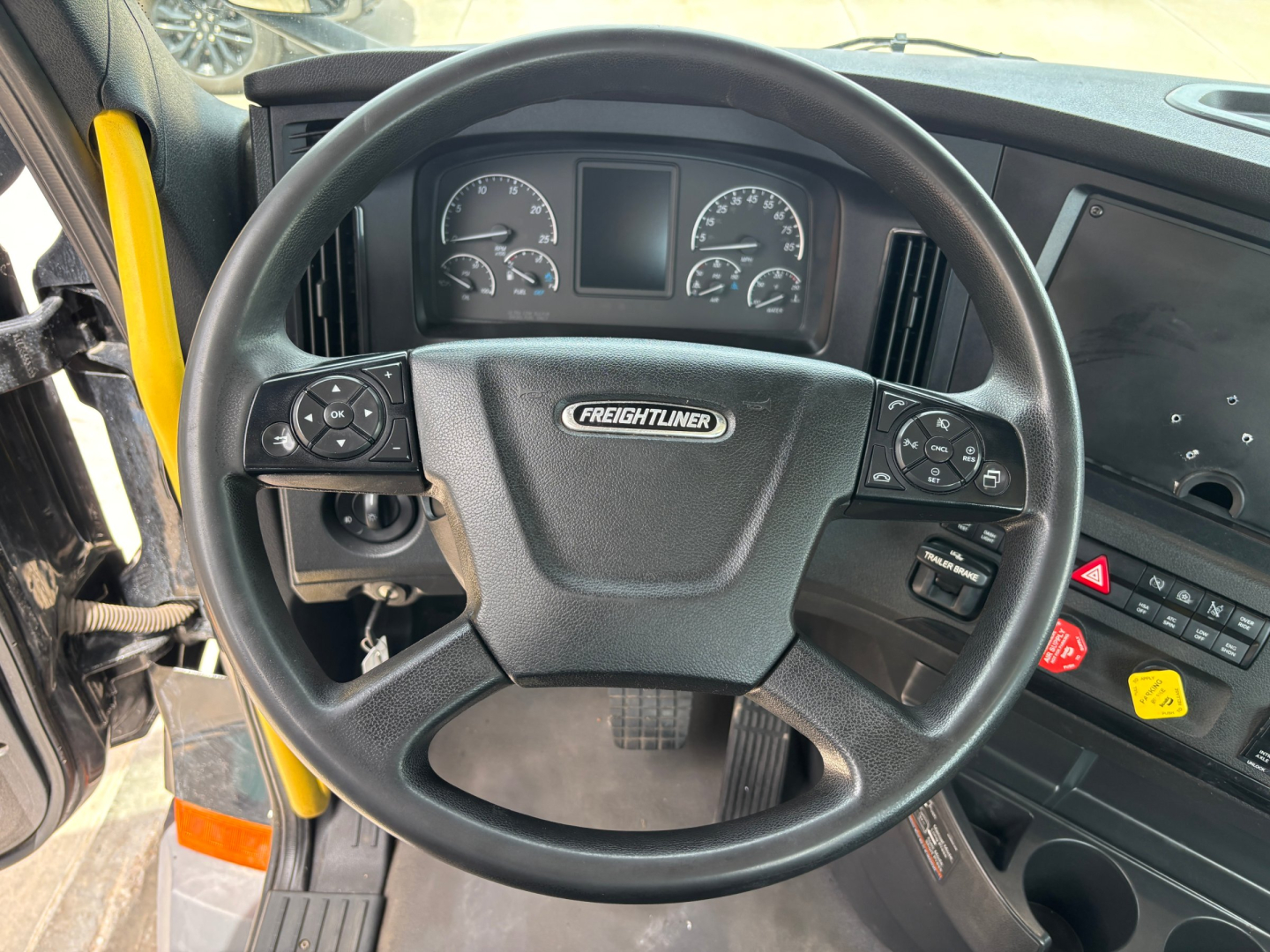 Freightliner steering wheel with dashboard and controls. Center badge reads Freightliner. Left and right control stalks on steering wheel, instrument cluster visible behind. Some dashboard buttons and hazard switch.