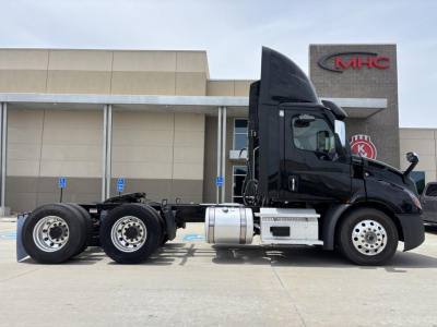 Truck side view showing 2023 Freightliner conventional daycab chassis with dual rear axles, black cab, fuel tanks, and exhaust, in a dealership lot.