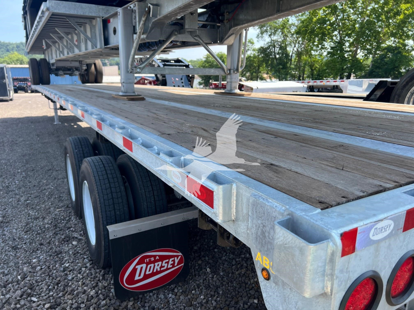 Flatbed trailer from rear-left showing metal frame, wooden deck, dual rear axles with tires, reflective strips, and Dorsey mudflap logo