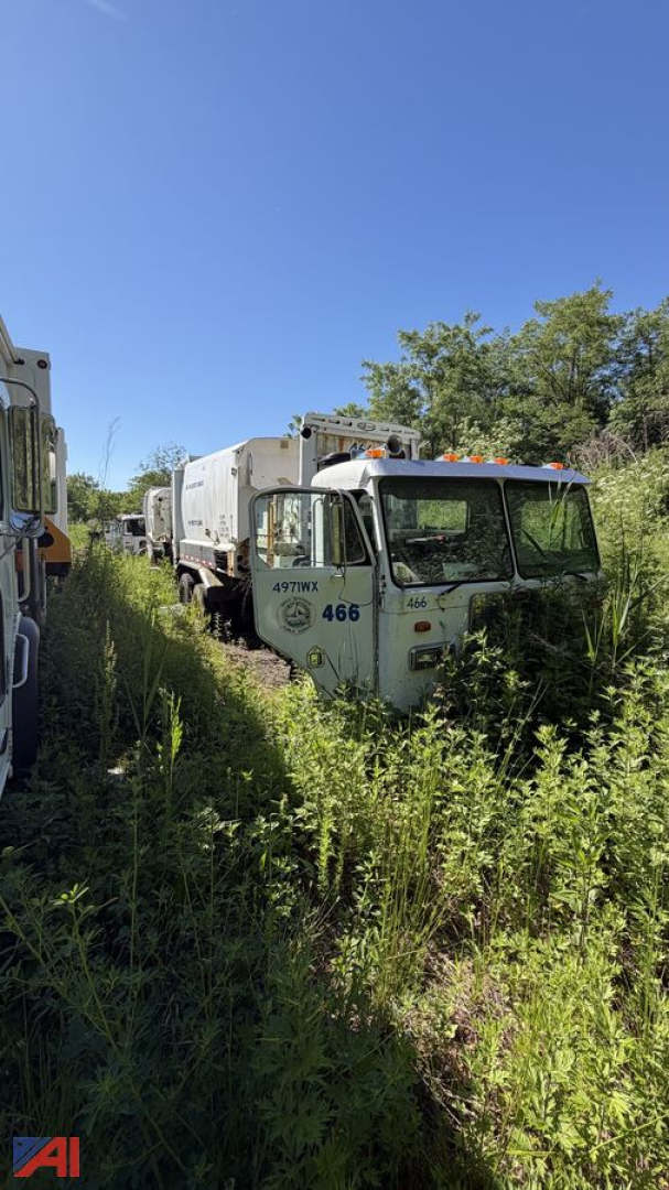 Peterbilt 320 garbage truck in overgrown lot viewed from front-left, with number 466 on cab and empty compactor body, other trucks in background.