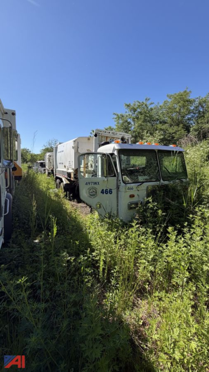 Peterbilt 320 garbage truck in overgrown lot viewed from front-left, with number 466 on cab and empty compactor body, other trucks in background.