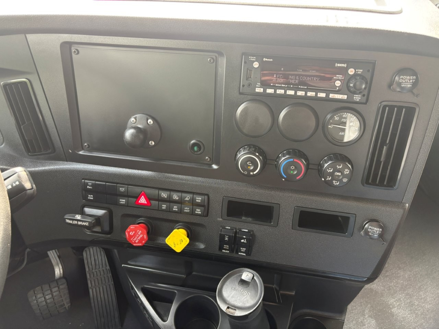 Cabin dashboard of a Freightliner Cascadia 126 showing gauge cluster, steering column area, air conditioning controls, hazard switch, two colored knobs, and radio unit.