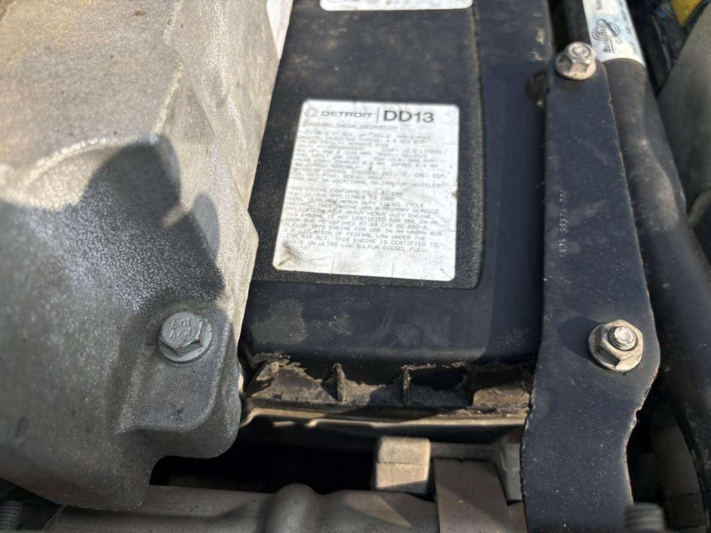 Label on a Freightliner engine bay with a DD13 label visible and surrounding components including bolts and a cracked plastic support.