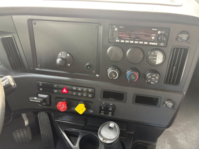 Cabin dashboard of a Freightliner Cascadia 126 showing gauge cluster, steering column area, air conditioning controls, hazard switch, two colored knobs, and radio unit.