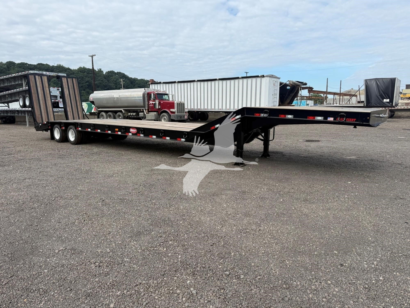 Lowboy trailer with a long flatbed showing black frame, dual rear axles, and gooseneck hitch; visible brand decals and reflective tape along sides.