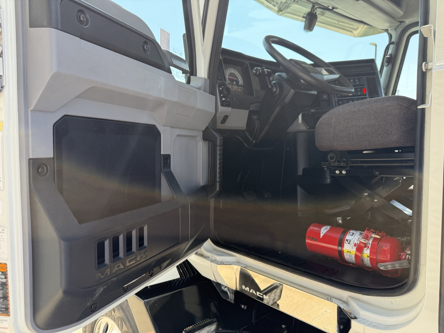 Cockpit and driver's door area of a 2021 Mack AN64T conventional daycab showing dashboard, steering wheel, and entry with a fire extinguisher mounted under the dash.