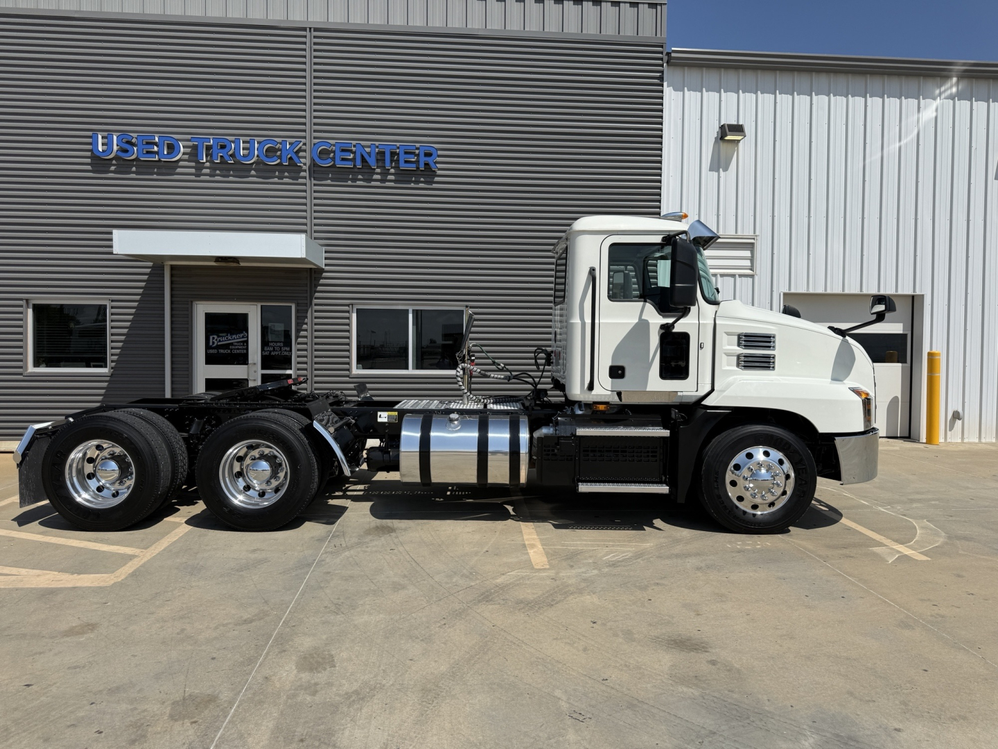Mack AN64T conventional daycab truck with white cab in front of used truck center building; visible VIN and stock number not in image; dual rear axles and chrome fuel tanks.