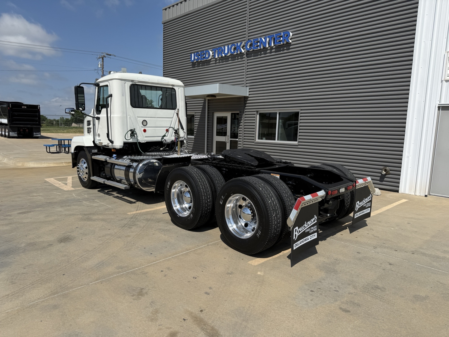 Mack AN64T daycab truck in front of a dealership, side view shows dual rear axles and fifth wheel, white cab, dealership building in background.