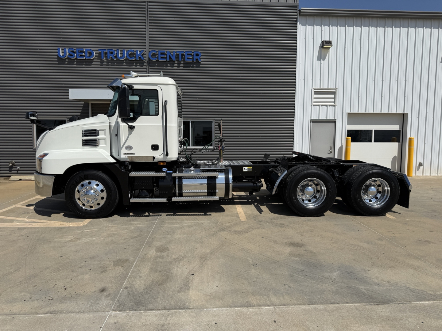 Truck chassis with cab and triple rear axles in a dealership lot, Mack AN64T badging not visible, white cab, flat concrete surface, building with USED TRUCK CENTER sign.