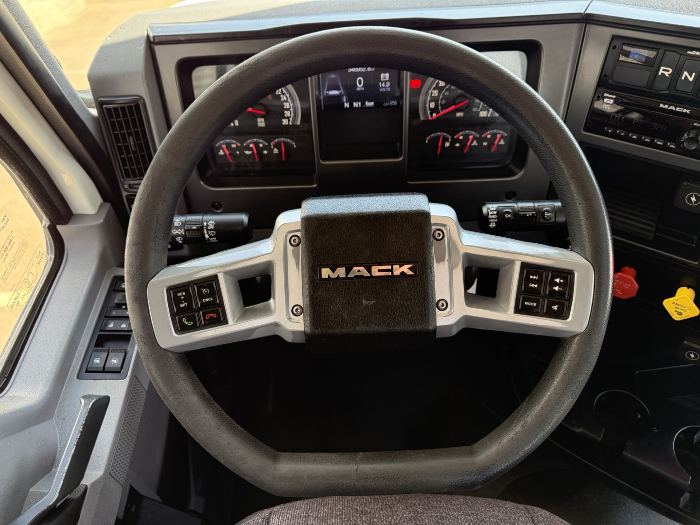 Steering wheel of a Mack truck dashboard showing controls and gauges, centered ‘MACK’ emblem on the wheel hub.