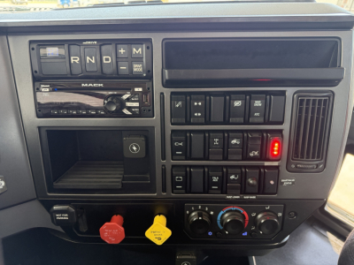 Truck dashboard showing gear selector with R N D + M, Mack stereo and controls, warning and climate dials, red and yellow buttons, and blank storage compartment.