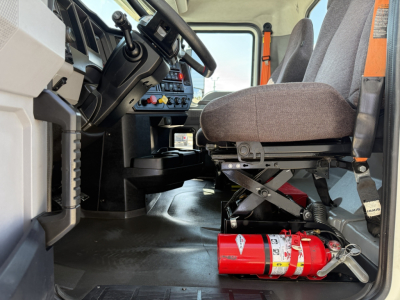 Truck cab interior showing steering wheel, dashboard, driver's seat, and a red fire extinguisher mounted beneath the seat in a Mack AN64T.