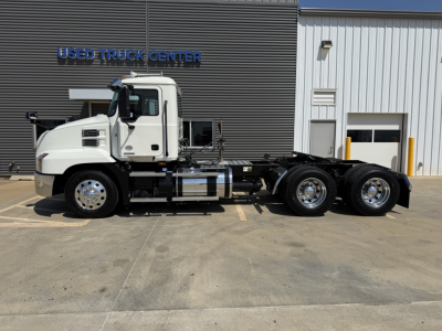 Truck chassis with cab and triple rear axles in a dealership lot, Mack AN64T badging not visible, white cab, flat concrete surface, building with USED TRUCK CENTER sign.