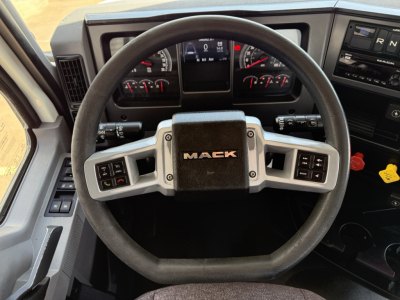 Steering wheel of a Mack truck dashboard showing controls and gauges, centered ‘MACK’ emblem on the wheel hub.