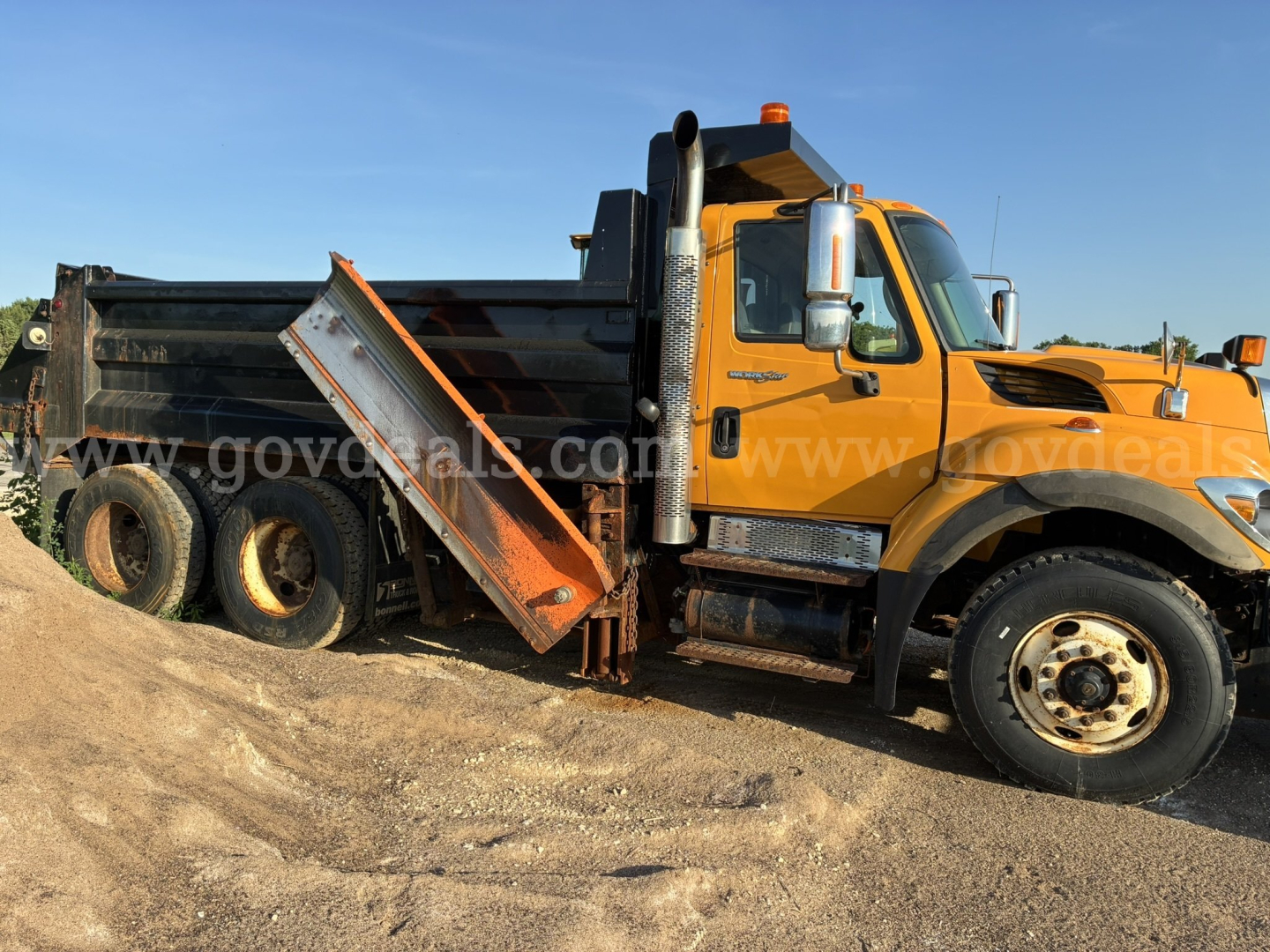 Dump truck side view showing hydraulic bed in raised position and black dump bed with orange cab, International logo visible, dual rear wheels.