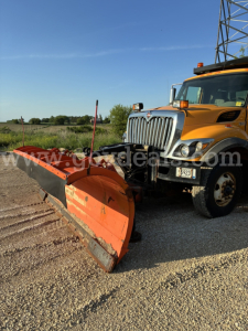 Orange snow plow attached to a yellow International truck in a gravel lot, plow blade visible with dirt and wear.