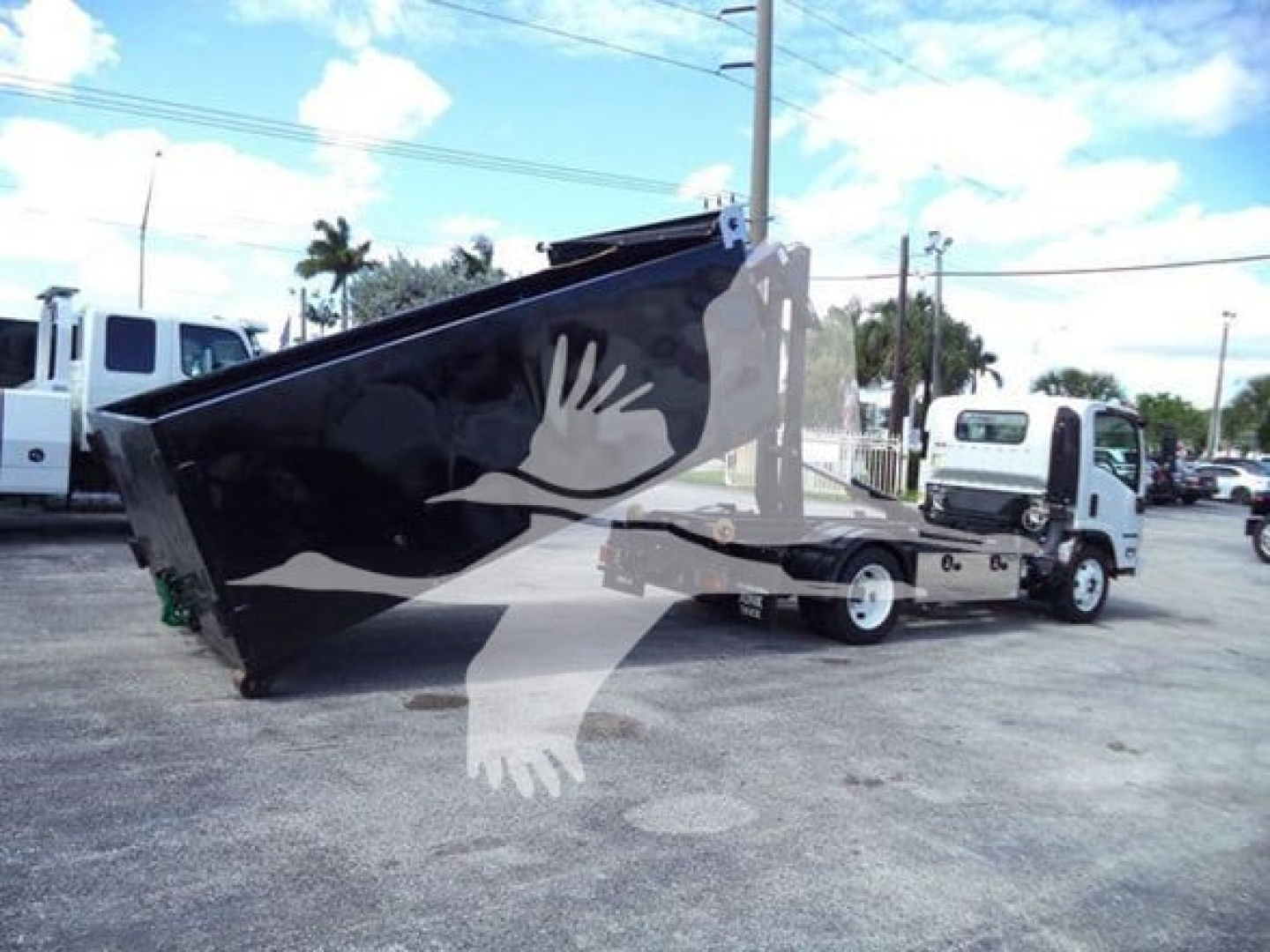 Isuzu NPR-HD truck with a large black hooklift bed, side view showing hoisted container and white cab, dealership lot in background.