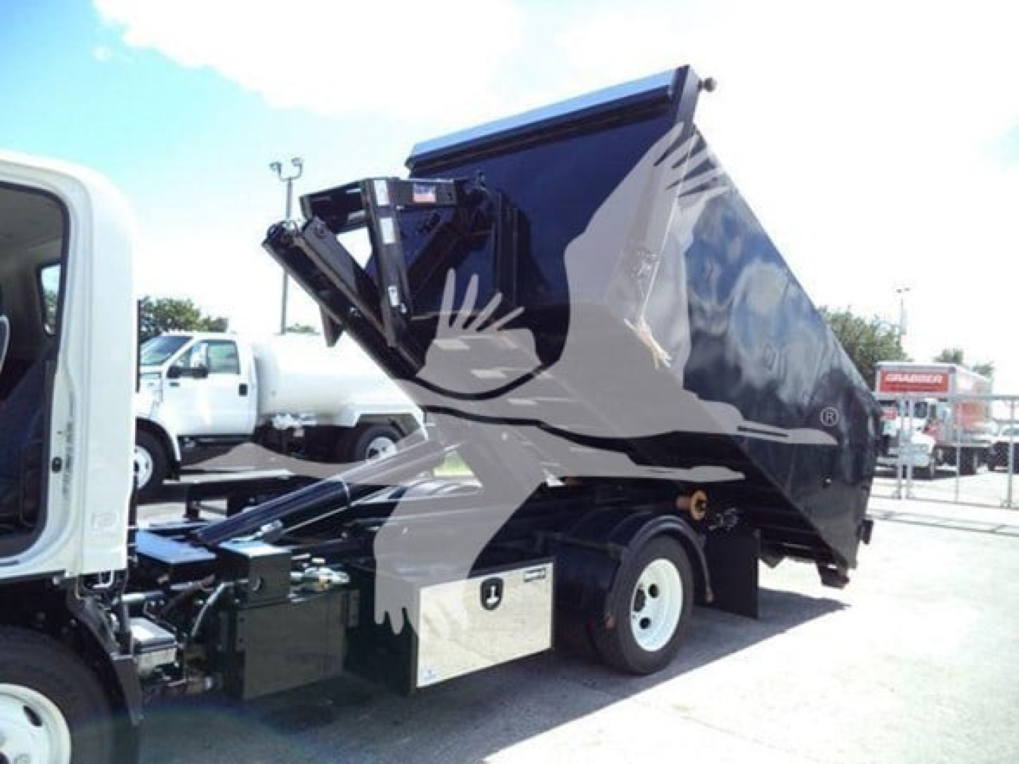 Isuzu NPR HD cab-forward truck with a raised hooklift system on the bed; side view showing a black hydraulic container and rear wheel; light blue sky, dealership lot in background.