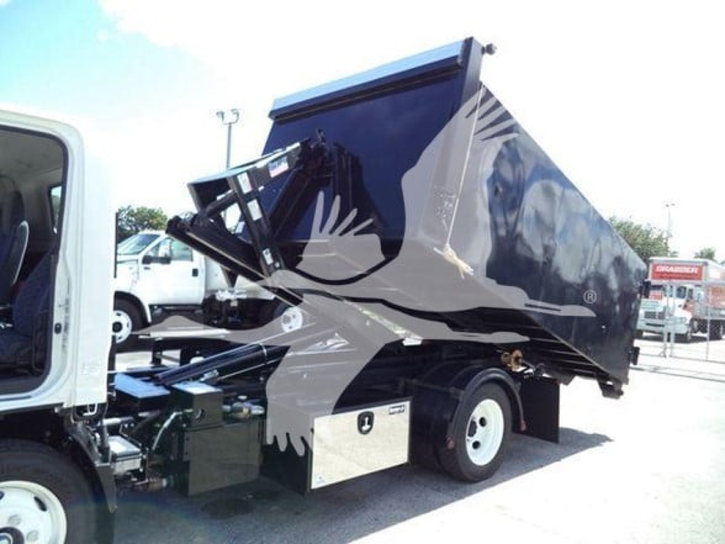 Isuzu NPR HD dump truck in a hooklift configuration with the bed raised. The image shows a black dumping bed on a white cab chassis, hydraulic mechanism visible. No legible serials or labels visible.
