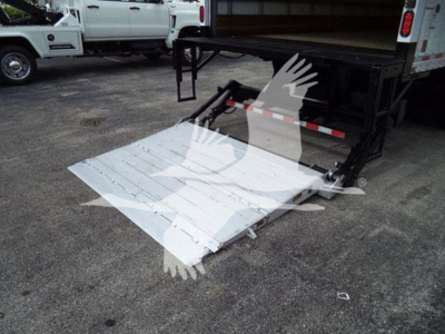 Truck bed with fold-out ramp on a white platform, attached to a trailer with hydraulic legs visible in background.