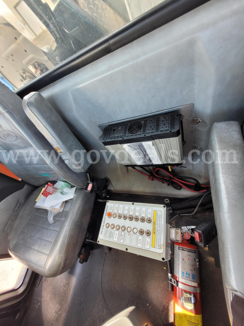 Vehicle cab interior with electrical components: a control/relay box with labeled switches and wiring, a power inverter module, and a mounted fire extinguisher visible near the seat, in a Mack GU713 truck cabin.