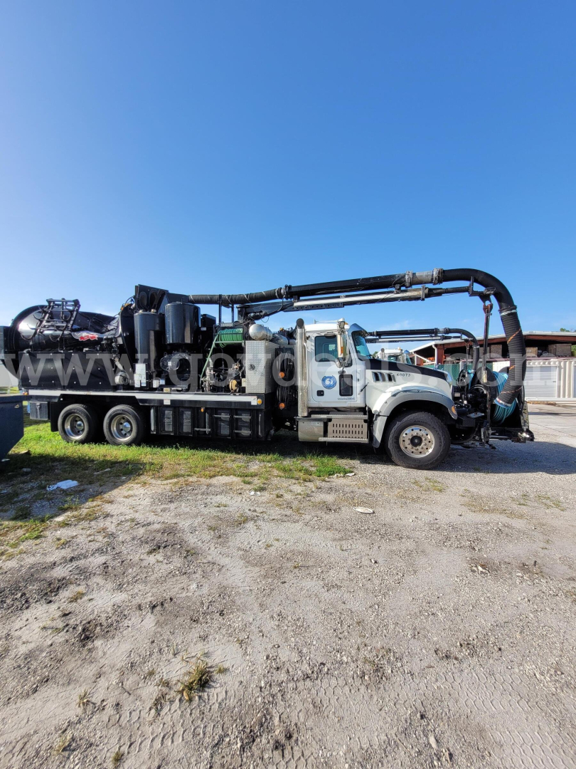 Truck-mounted vacuum unit on a Mack GU713 with large black hose reel and crane-like piping along the chassis, front cab visible, clear blue sky.