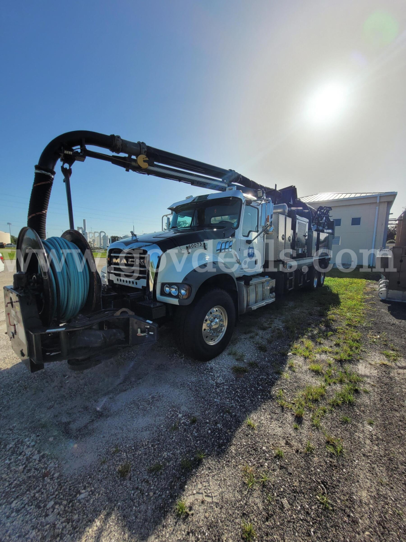 Truck-mounted vacuum unit showing a large hose reel and crane rig on a white Mack GU713 chassis.