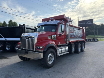 A red Western Star dump truck in a dealership lot with a chrome front grille and dual rear axles; visible text includes 'Western Star' on the sign in the background and on the truck's badge.