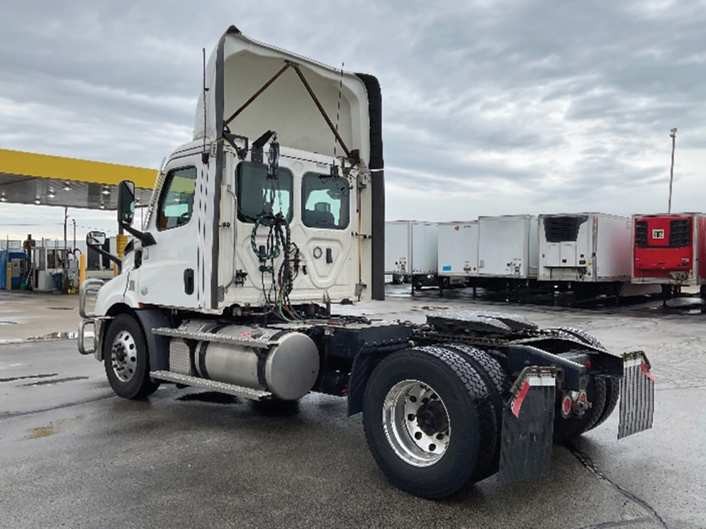 Truck chassis with cab tilt showing Freightliner Cascadia 116 tag, side-mounted fuel tanks, rear axle, and exposed wiring; daytime outdoor lot
