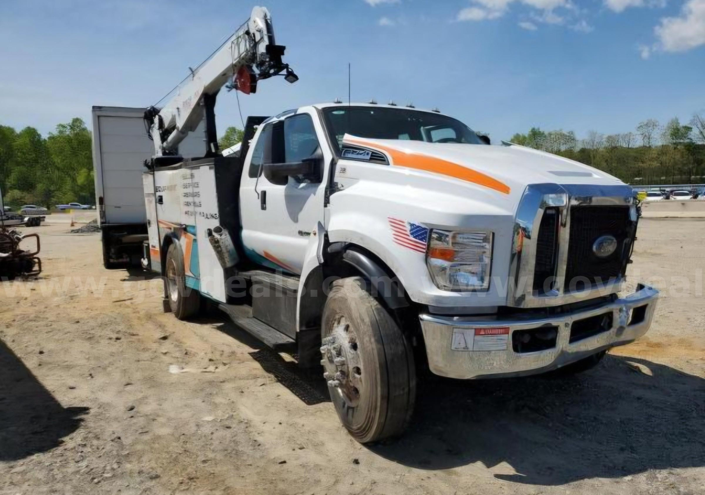 Ford F-750 tow truck with crane, front grille and headlights visible; service/utility body with orange stripe; American flag decal on the door; wheel and tire shown; stock number and service truck branding may be on the service body.