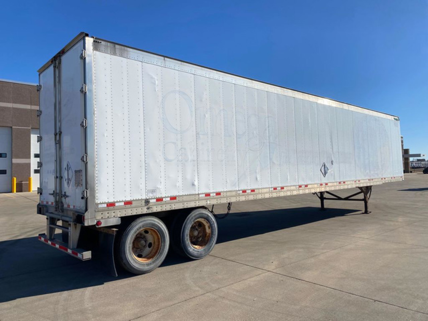 Trailer side view showing two rear wheels and metal freight trailer with diamond-shaped hazard placard area, no visible branding text legible.